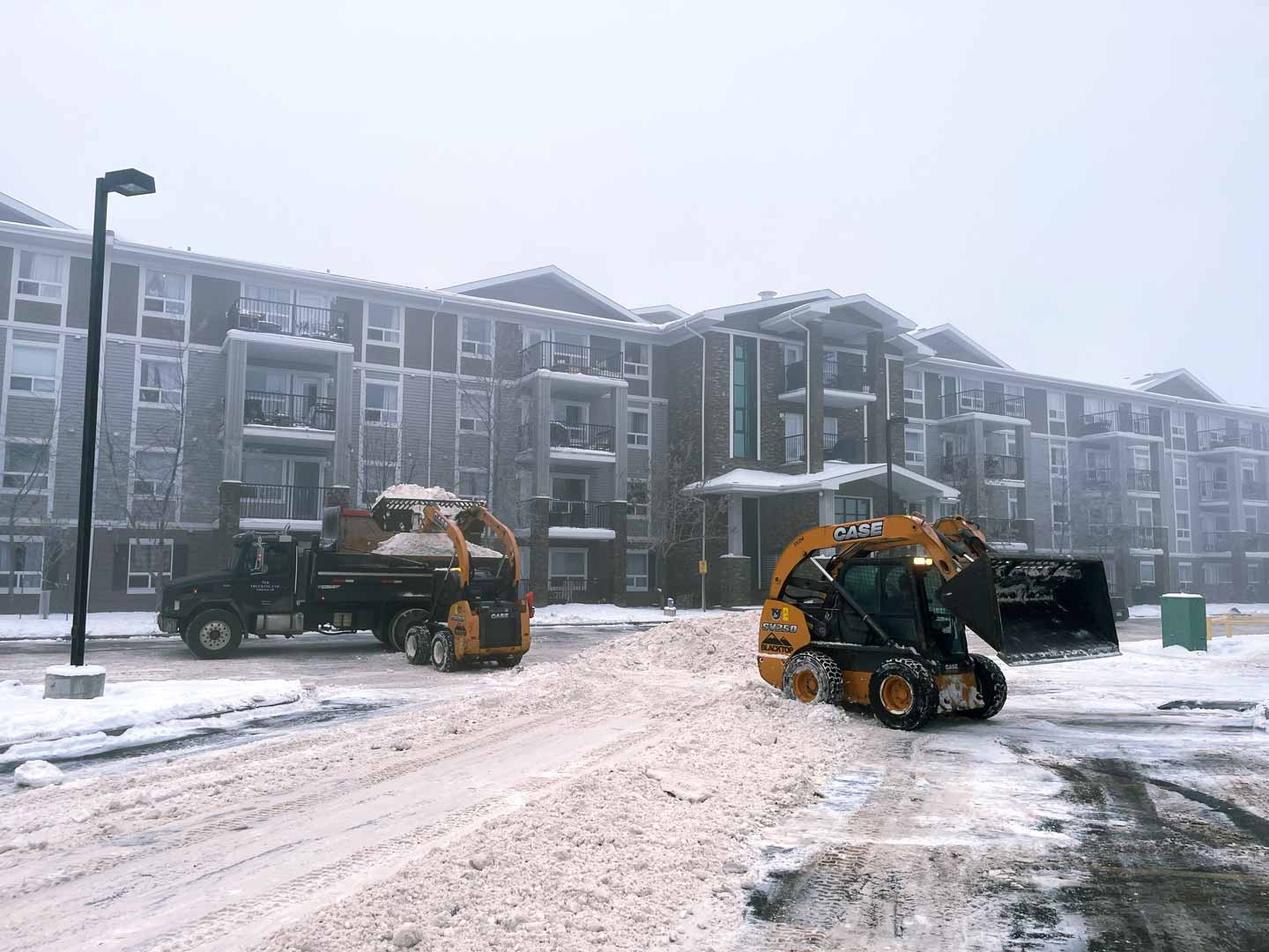 Skid steers removing snow and hauling snow away in parking lot complex in Edmonton Alberta.